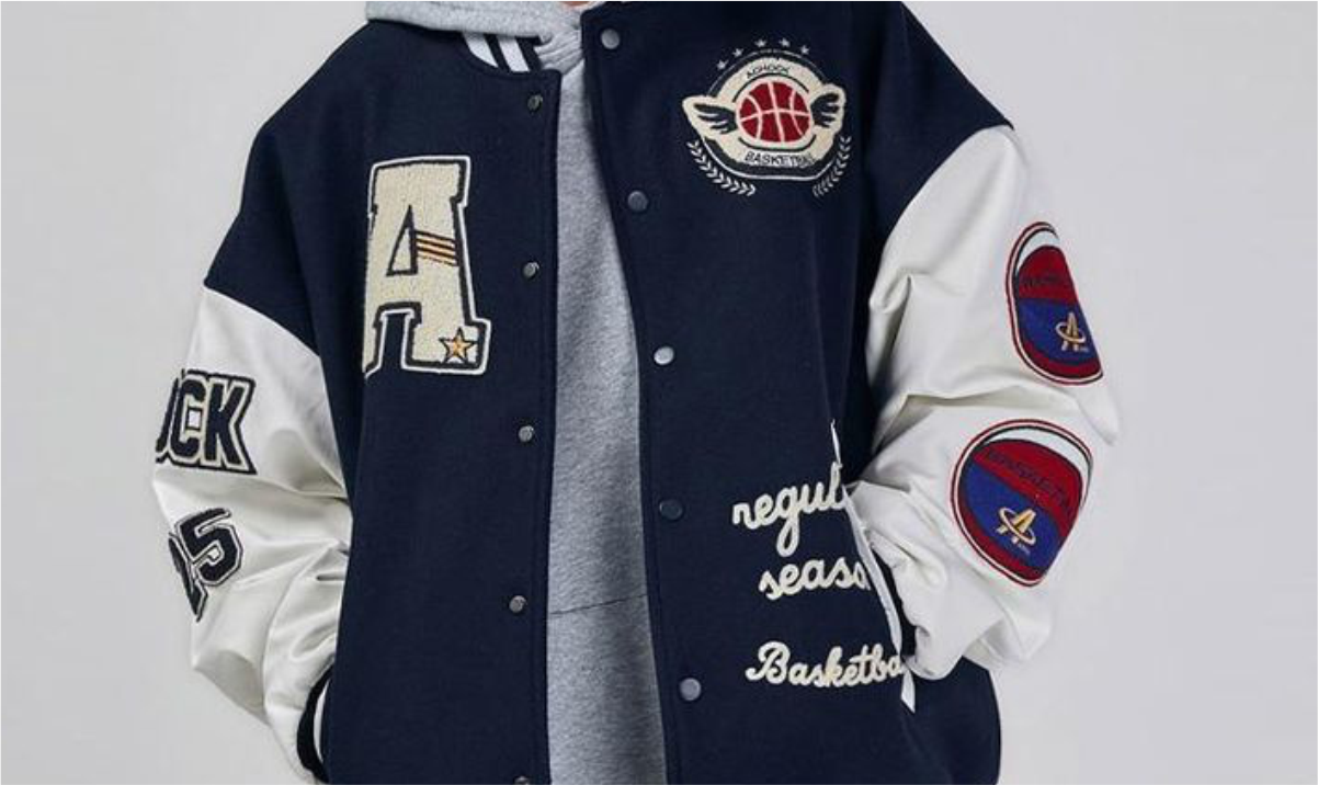 Jaket varsity biru navy berlengan krem kontras dipakai seseorang, terlihat kancing snap logam, rib kerah dan pergelangan bergaris, serta patch huruf chenille di dada