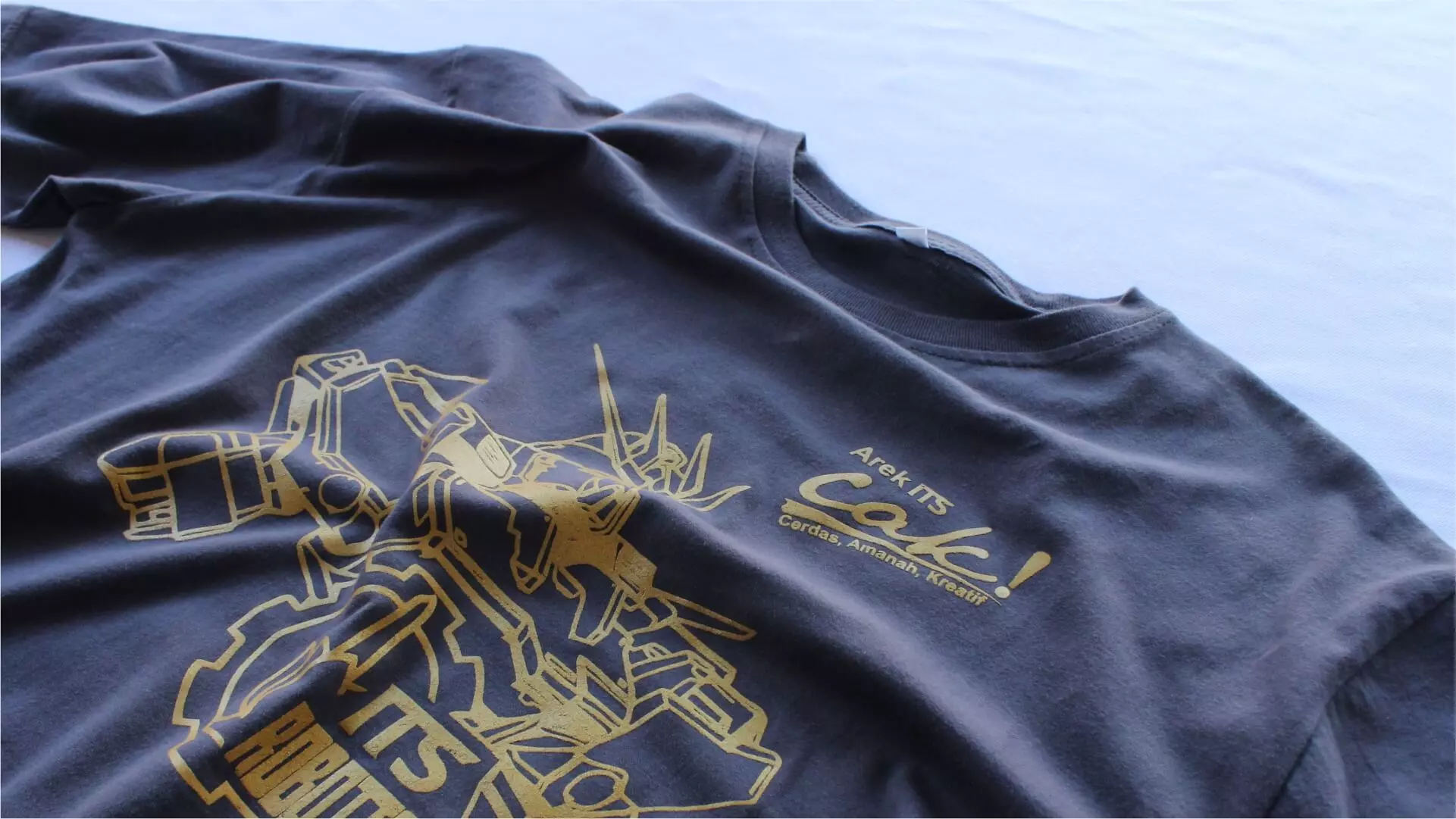 Kaos abu-abu bersablon emas tergeletak rata di atas kain biru muda