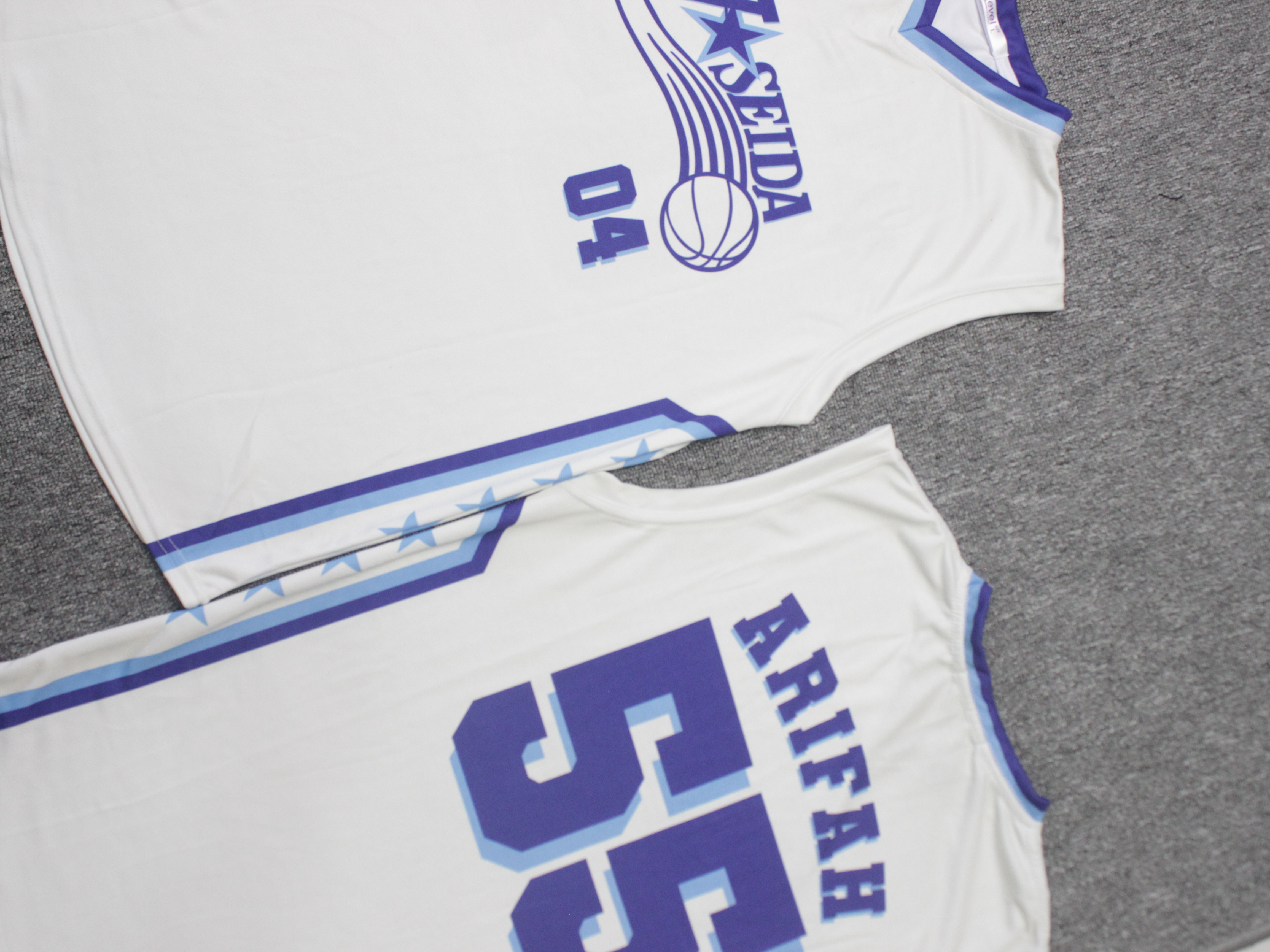 Dua jersey basket custom dengan nama dan nomor sublimasi tergeletak di karpet produksi