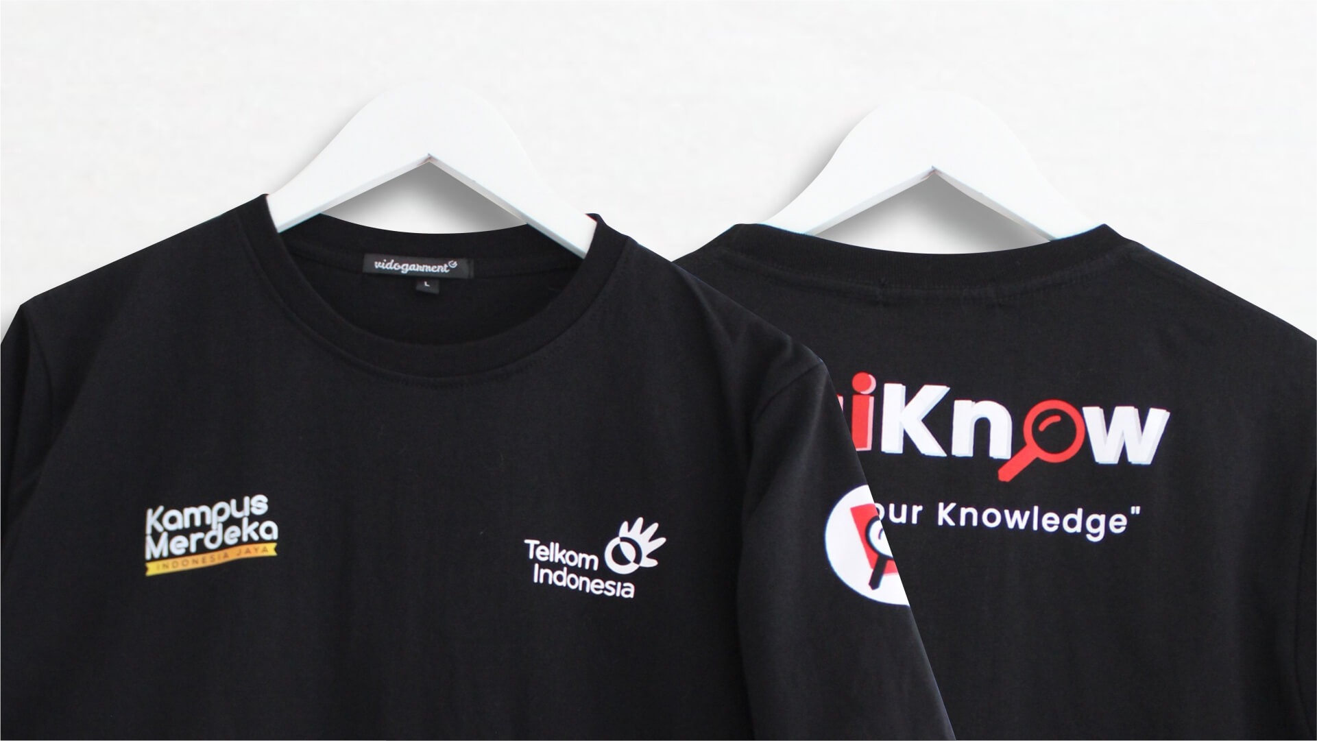 Dua kaos hitam bersablon tergantung di hanger, satu dengan cetakan punggung besar putih beraksen merah