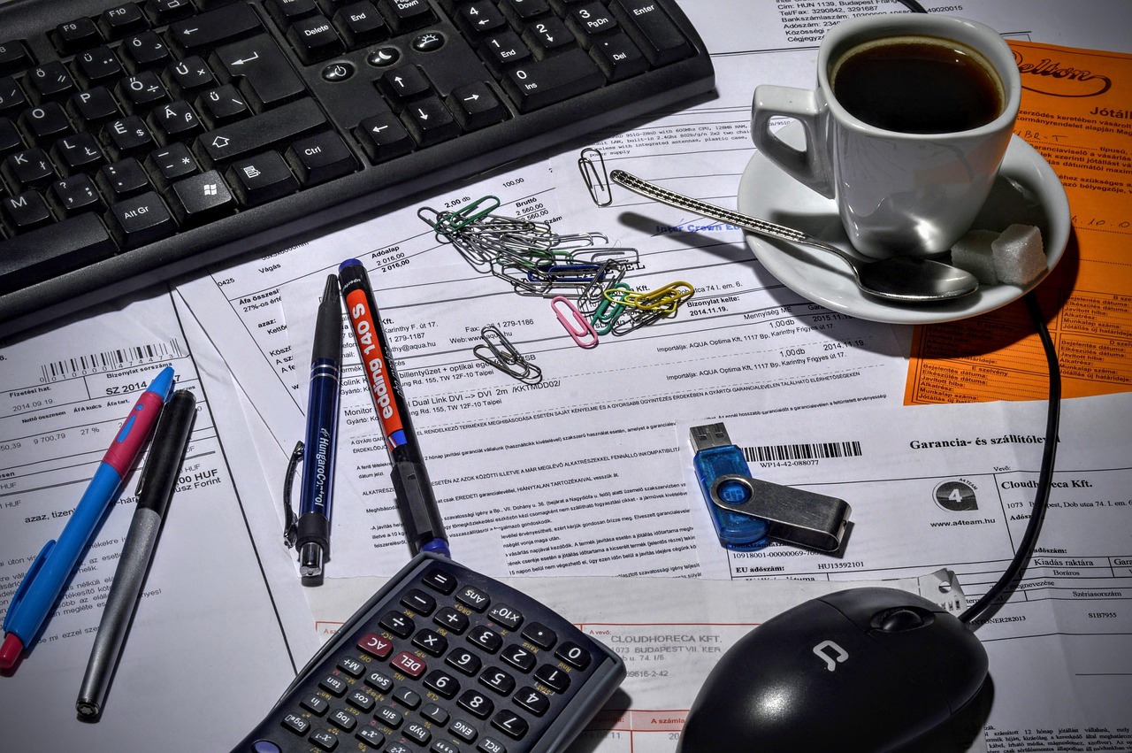 Meja administrasi dari atas: tumpukan tagihan dan kertas kerja dengan kalkulator, pulpen, paperclip, dan cangkir kopi di sekelilingnya