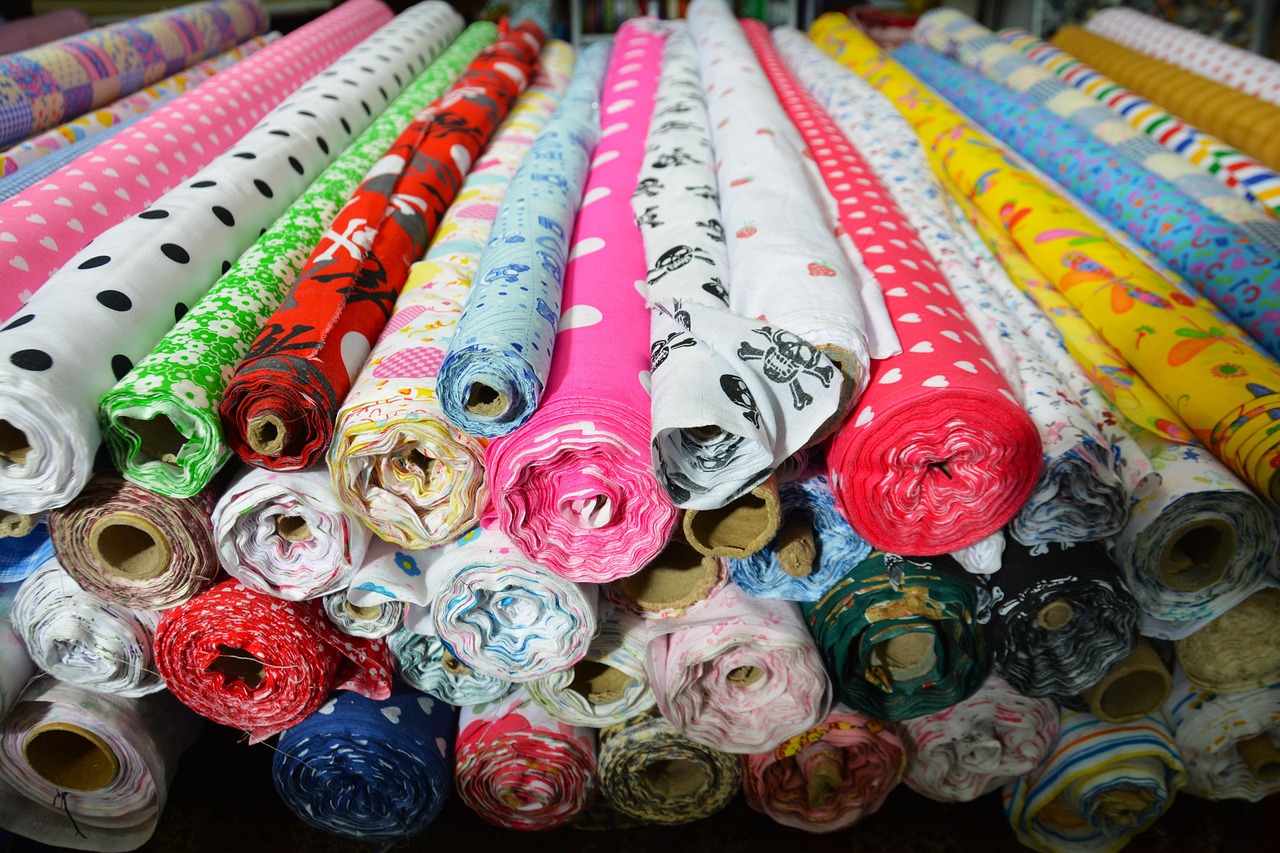 Tumpukan gulungan kain beragam warna dan motif, termasuk beberapa gulungan putih, bersusun di toko kain
