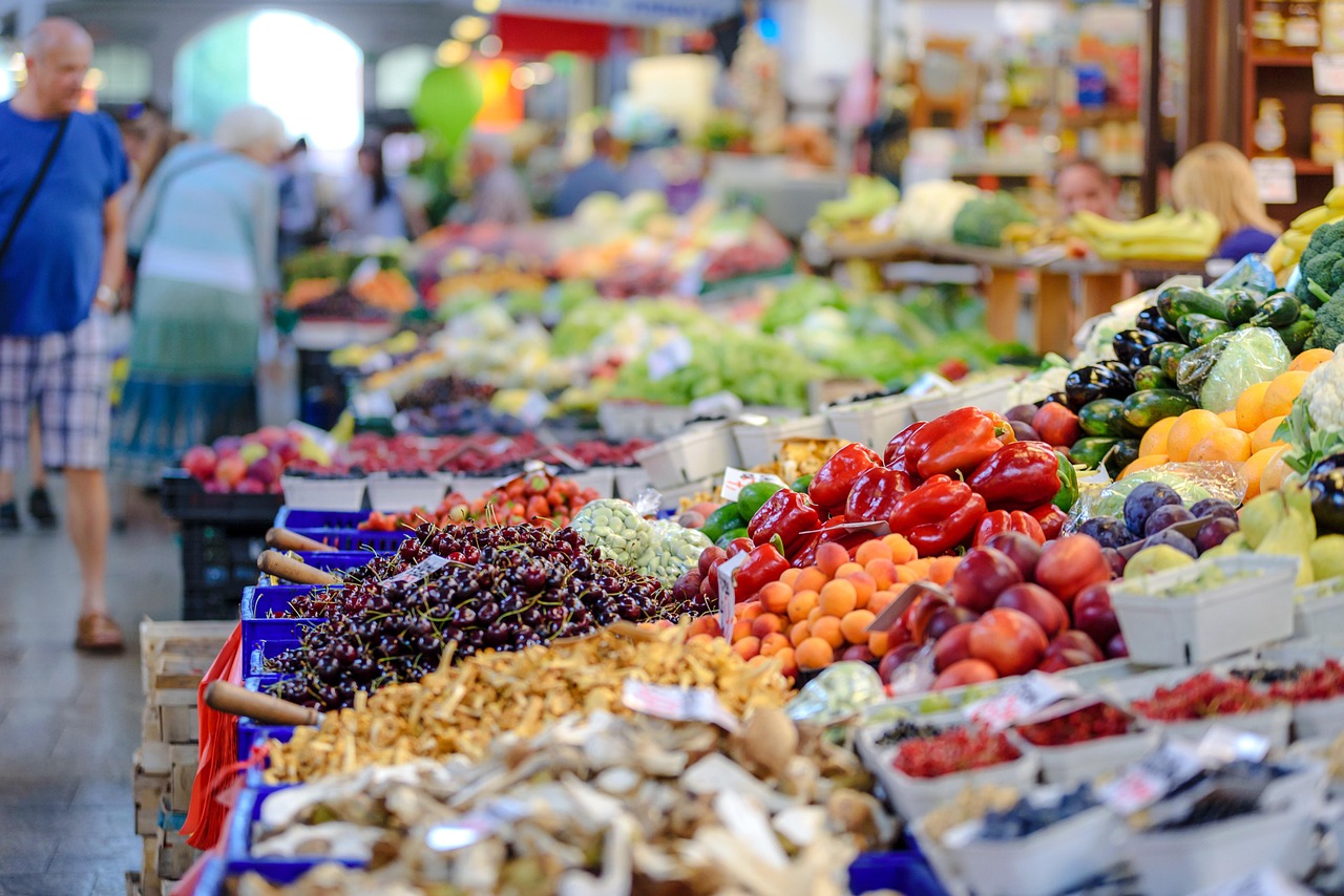 Buah dan sayur bertumpuk di meja kios pasar, seorang pembeli memeriksa dagangan di belakangnya