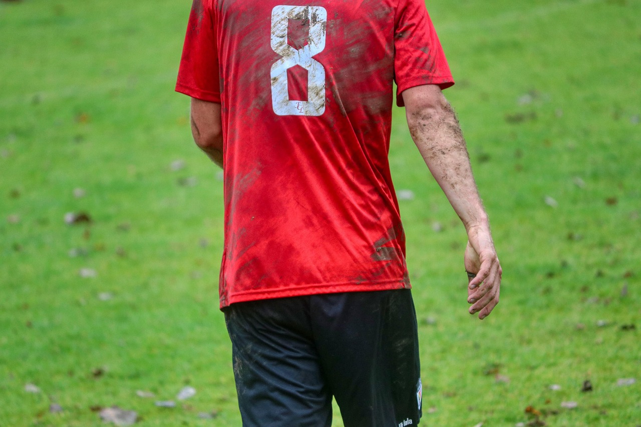 Punggung jersey merah bernoda lumpur dengan nomor putih 8 yang tercetak menyatu dengan kain, difoto dari belakang di lapangan rumput