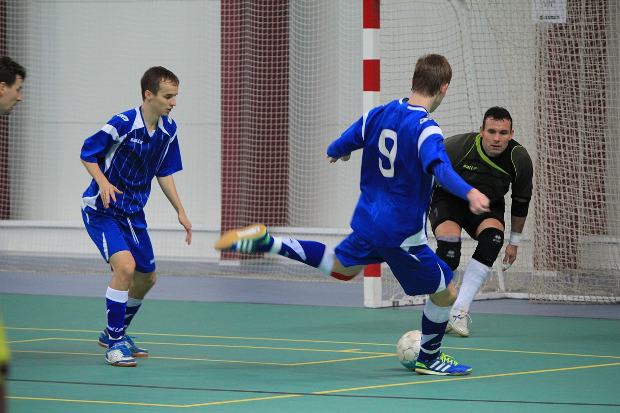 Pemain futsal dengan set seragam biru dari atasan, celana, sampai kaus kaki menendang bola di dalam gedung olahraga; nomor punggung 9 terlihat di punggungnya